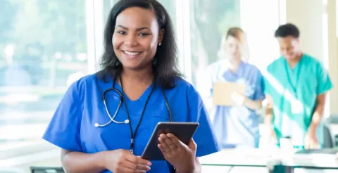 Smiling nurse in blue scrubs holds a tablet and wears a stethoscope, while two healthcare workers confer in a bright hospital setting.